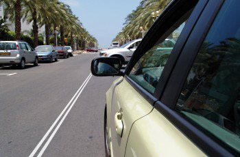 מבט ממראת הרכב על כביש עם קו הפרדה רצוף וחניית רכבים משמאל תחת עצי דקל.