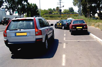 חניון עם רכב כסוף נוסע בשביל, ורכב כהה יוצא מחניה לצדו.