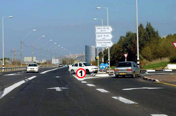 צומת עם רכב לבן שמתחיל לפנות שמאלה מול הרכב שלך, ושלטי הכוונה בצד הדרך.