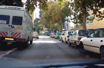 דרך עירונית צרה עם רכבים חונים בצפיפות משני צדי הכביש, ובהם רכב שירות עם לוחות אזהרה אדום-לבן בצד שמאל.