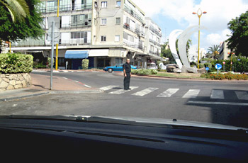 גבר עומד בתוך מעבר חצייה מסומן בכיכר עירונית פתוחה, ליד פסל לבן גדול ורחוב מסחרי ברקע.