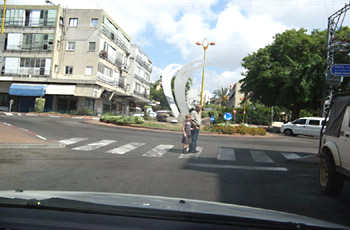 שני הולכי רגל חוצים מעבר חצייה מסומן בכיכר עירונית, ליד פסל לבן גדול.