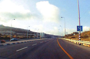 תמרור כחול מלבני על עמוד גבוה בצד ימין של כביש מהיר מרובה נתיבים, עם מעקה בטיחות לאורך השול.
