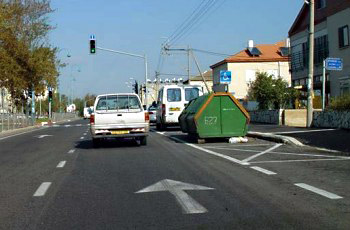 צומת עם רמזור, מכולת אשפה ירוקה ורכב מסחרי לבן חונים בצד הדרך, חץ צהוב על הכביש