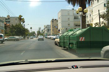 רחוב עירוני עם מכולת אשפה ירוקה גדולה החוסמת חלק מהמדרכה בצד ימין